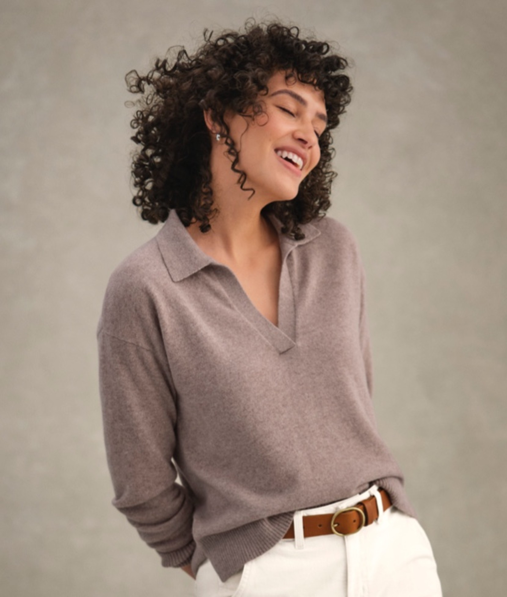 A woman in a mauve collared sweater tucked into white pants, standing.