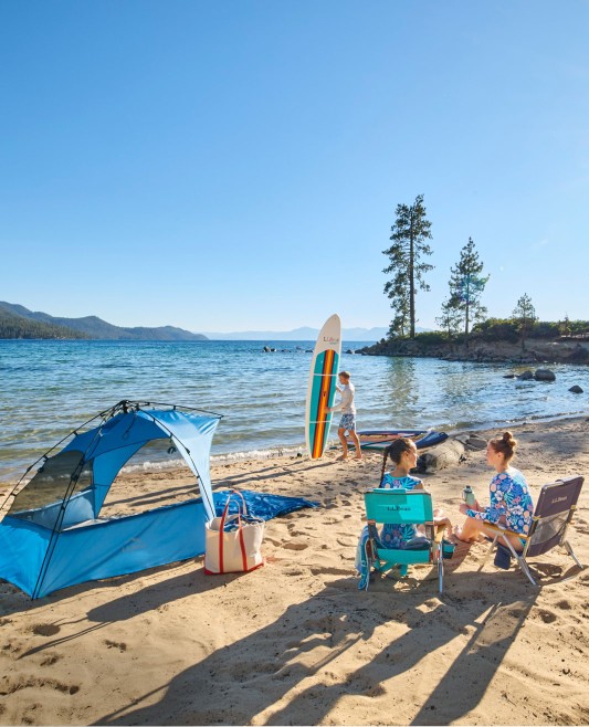 3 people enjoying a day at the beach, 2 women on beach chairs and a man with a stand-up paddleboard.
