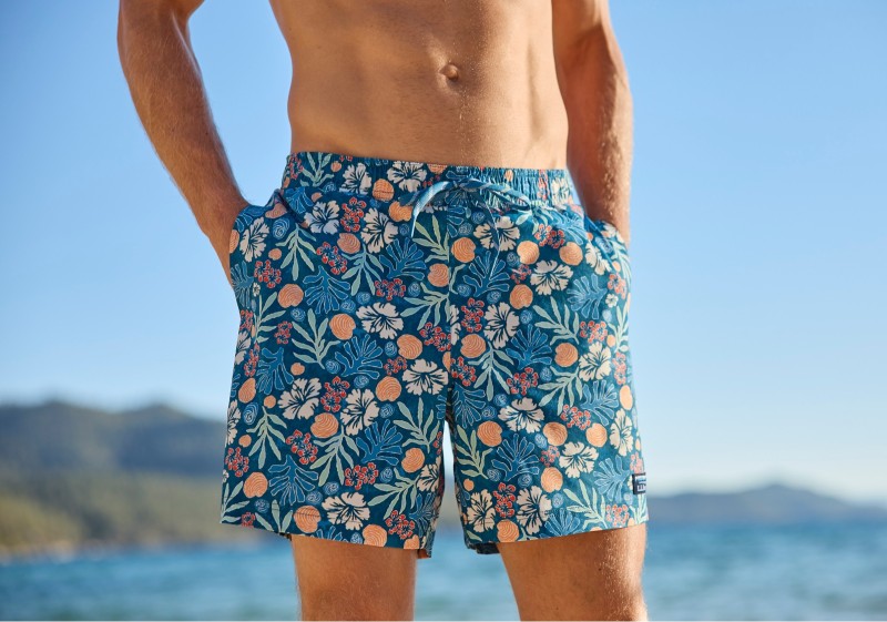 Close up of a man knee to chest wearing swim trunks, ocean in the background.
