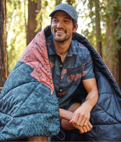 Close-up of man sitting in a forest with a blanket around his shoulders.