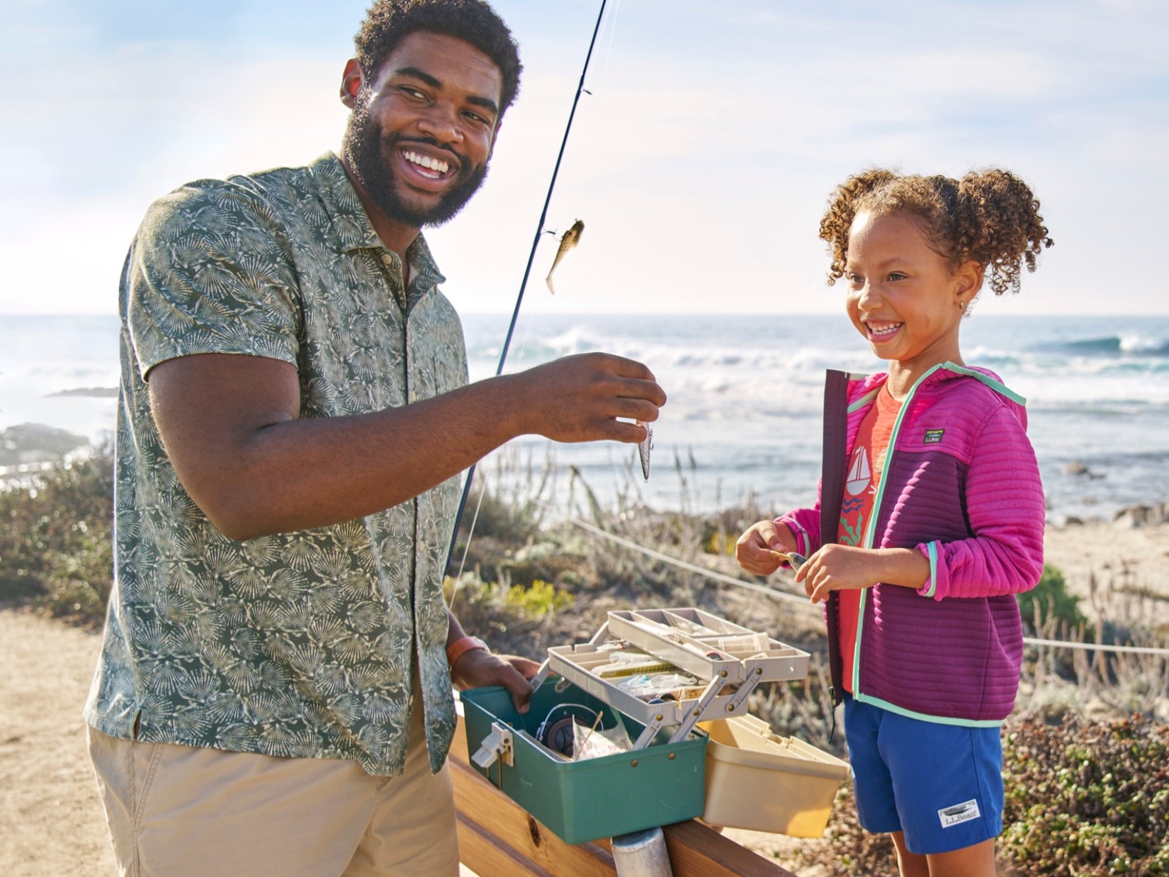 A man on the beach holding a fishing rod and open tackle box showing a young girl a fishing lure.