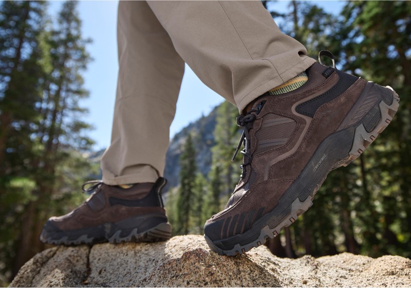 Close-up of hiking boots being worn by someone walking on a rocky surface, mountains in the background.