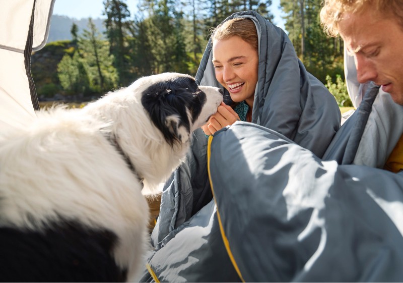 Man and woman wrapped in sleeping bags, sitting on the ground outside with a dog.