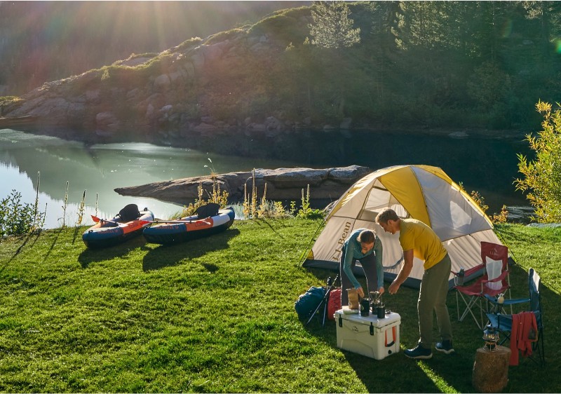 A campsite by the water with tent, camp chairs and 2 people making coffee.