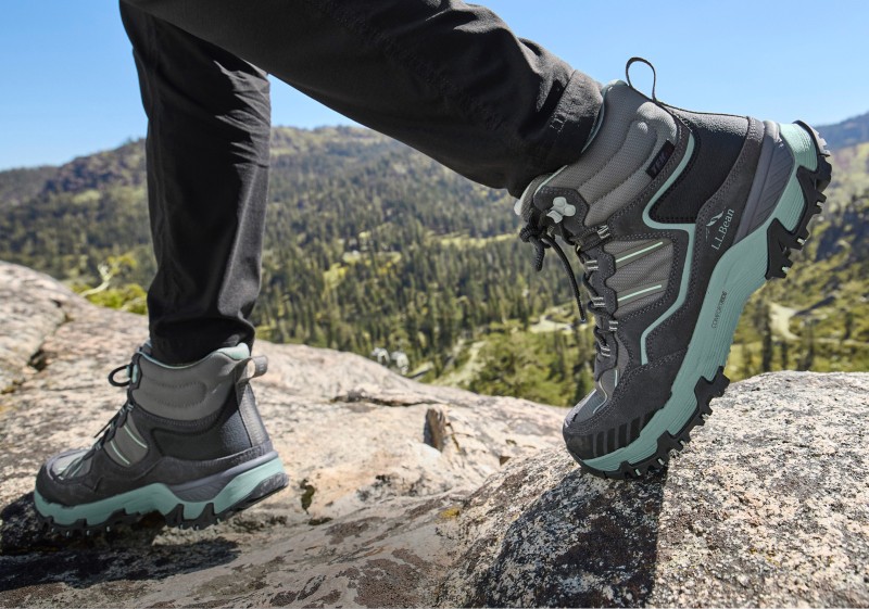 Close-up of hiking boots being worn by someone walking on a rocky surface, mountains in the background.
