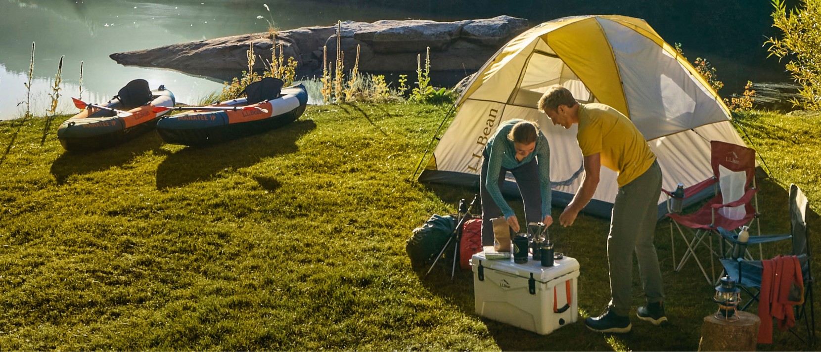 Two people making coffee at a beautiful campsite by the water with a tent, camp chairs and kayaks.