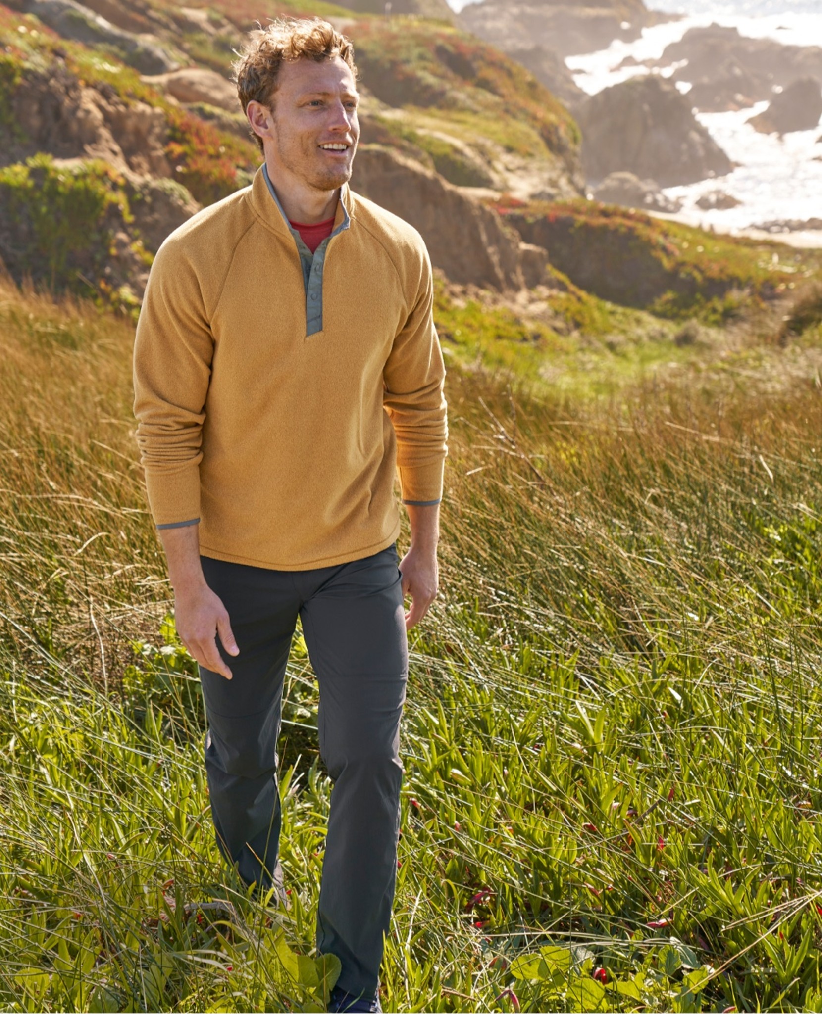 Man in yellow sweater walks through tall grass near rocky cliffs and ocean.