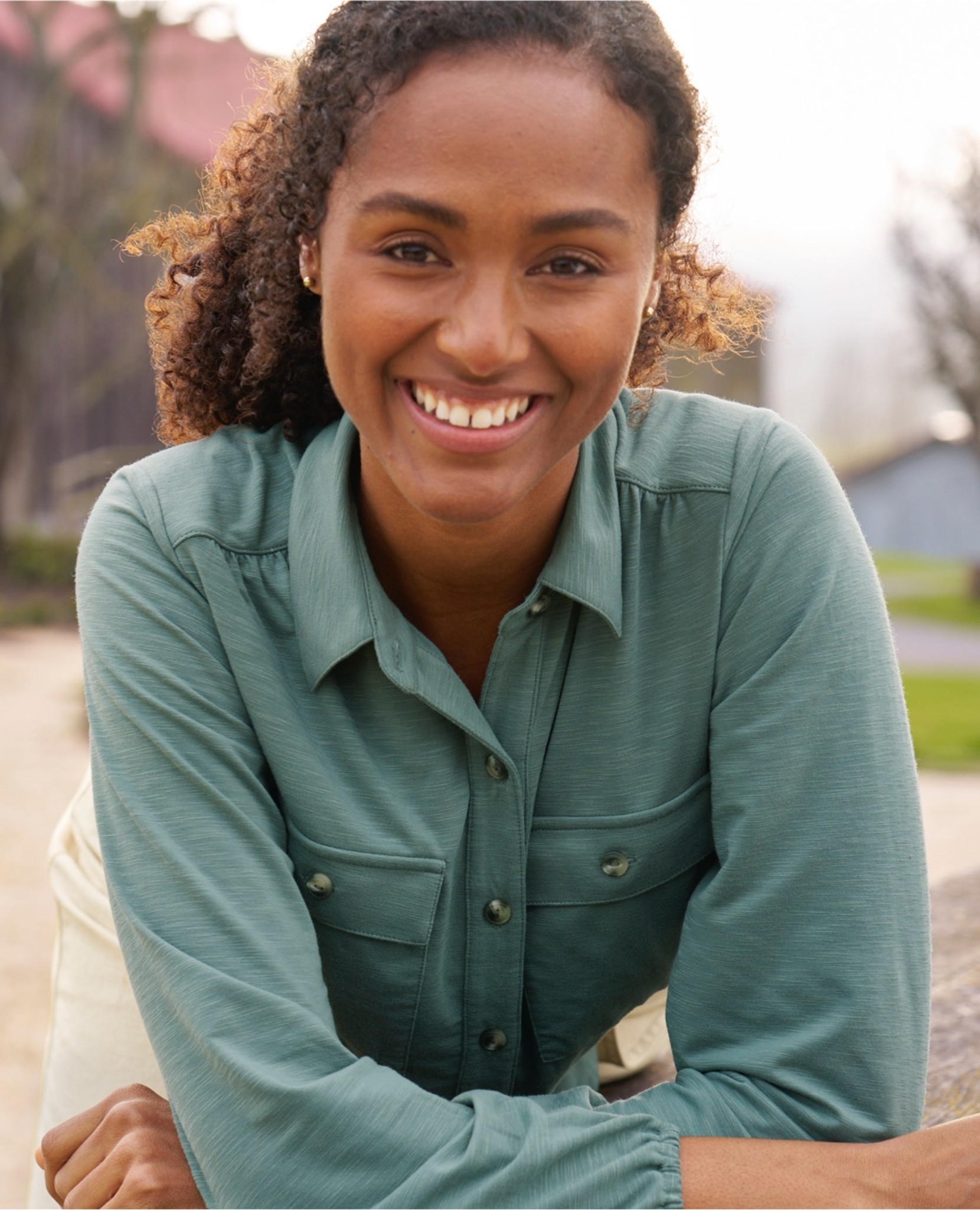 Woman with curly hair in a green shirt leans forward with arms on a surface; outdoor background visible.