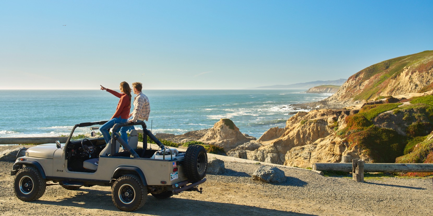 Two people sit on a Jeep by a coastal cliff, one pointing toward the ocean under a clear blue sky.