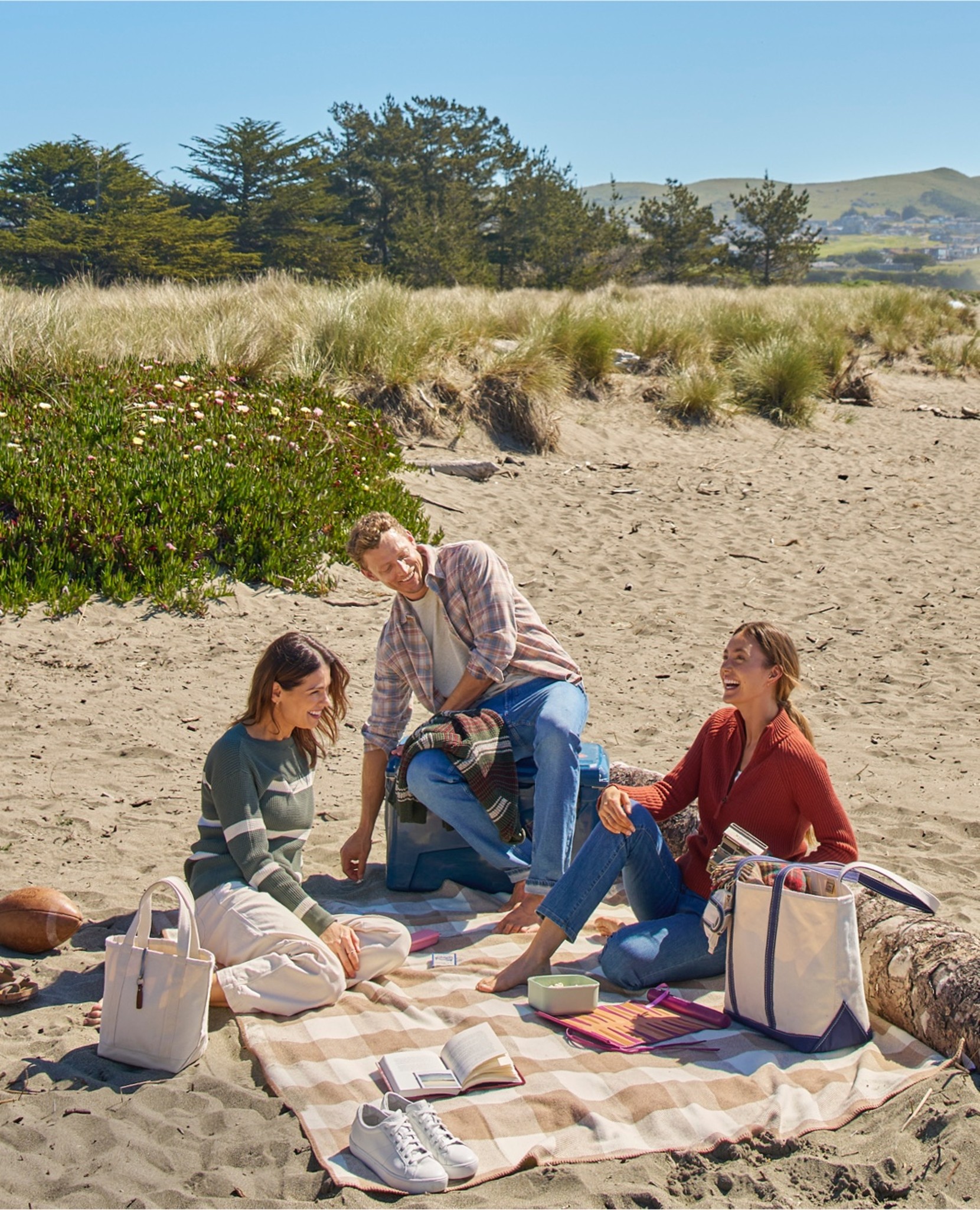 Three people sit on a blanket at the beach with bags and a football nearby; grassy dunes and trees are in the background.