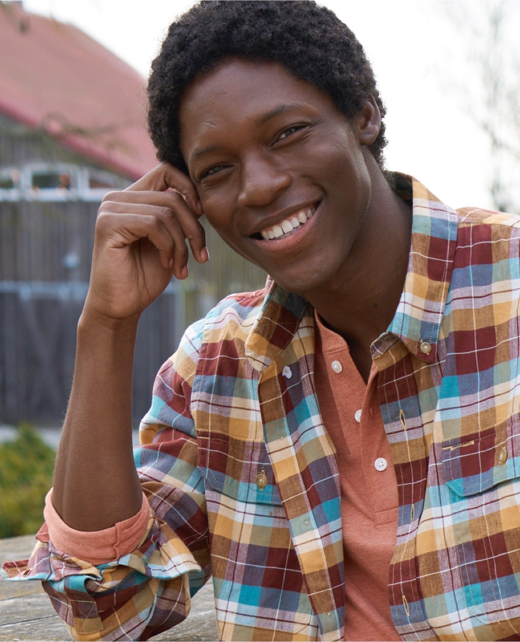 Man in a colorful plaid shirt over orange henley sits outdoors with hand on head.
