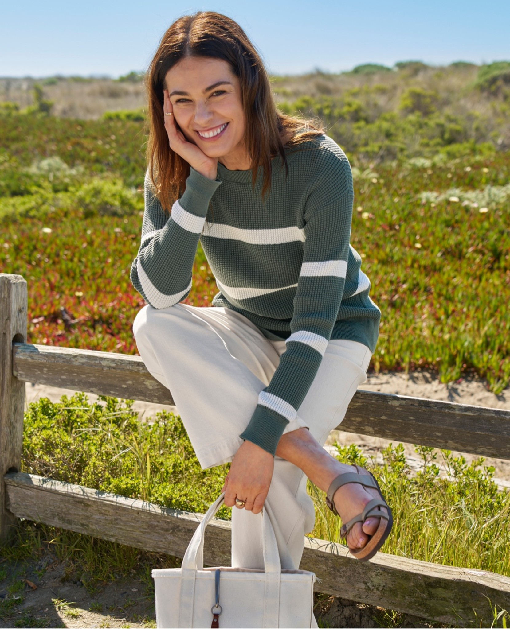 Woman in striped sweater and white pants sits on a wooden fence outdoors, holding a white tote bag.