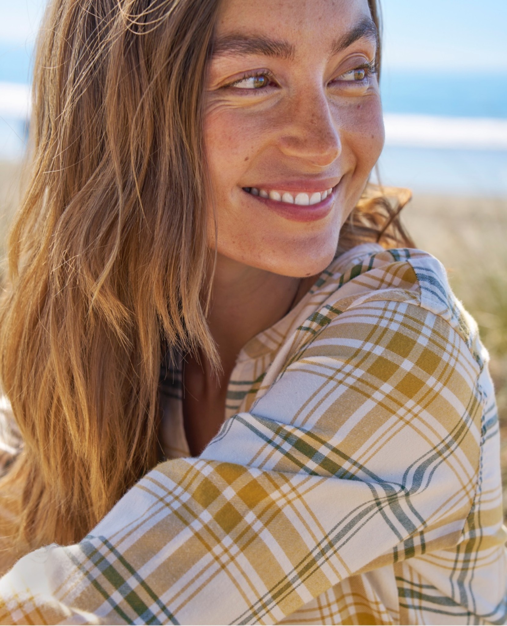 Woman with long blonde hair in a yellow plaid shirt sits outdoors near the beach.