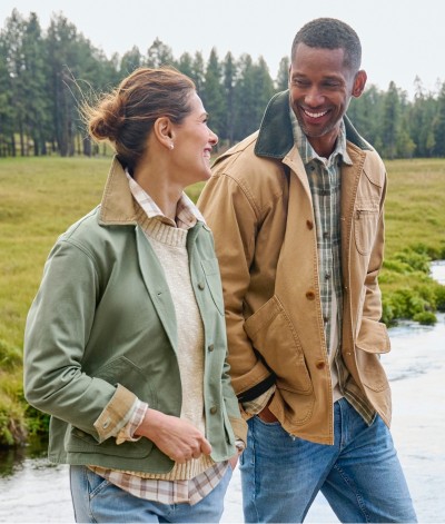 Two people in jackets and jeans walk near a river, smiling at each other with trees in the background.