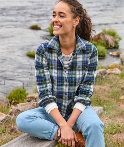 Woman in blue-green plaid shirt sits smiling on a riverside bench with rocks, grass, and water in the background.