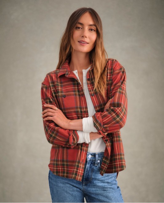 Person in red plaid shirt over white top and jeans stands with arms crossed against a neutral background.
