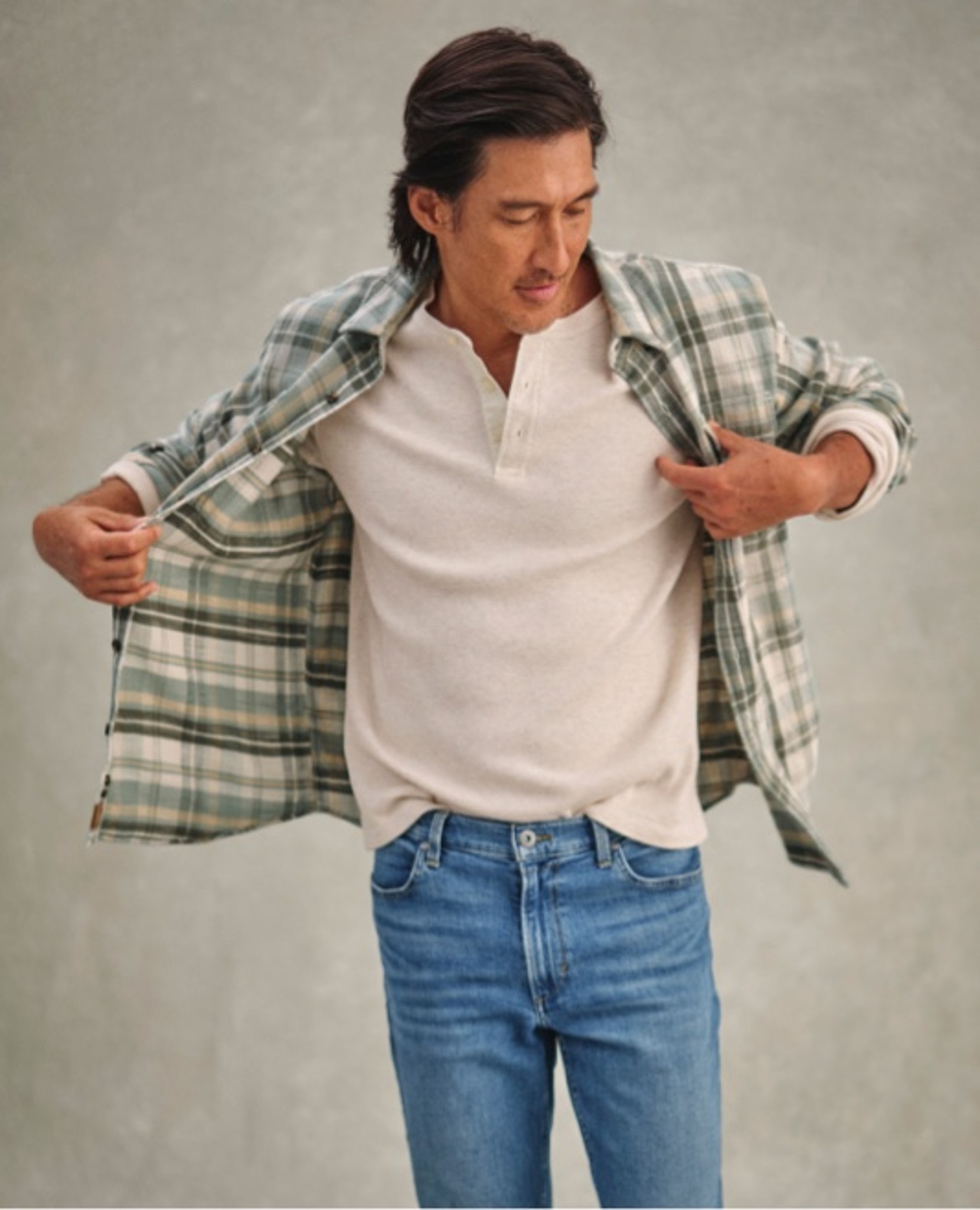 Person adjusts green plaid shirt over white henley and jeans.