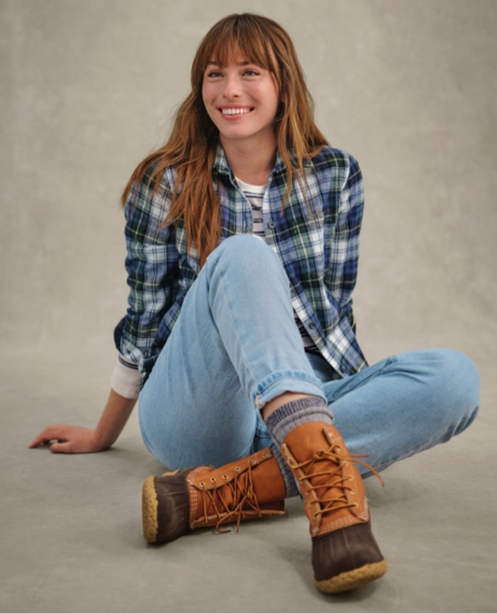 Person sits on floor in plaid shirt, striped top, jeans, and boots.