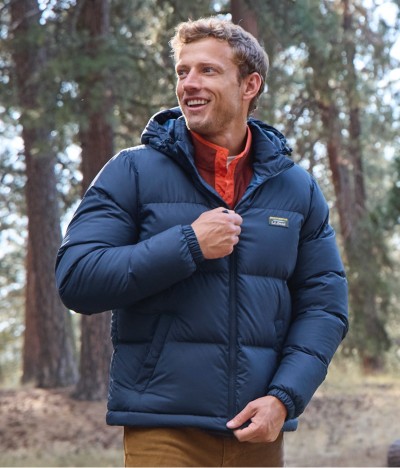 Man zipping blue jacket with orange scarf, standing among trees.