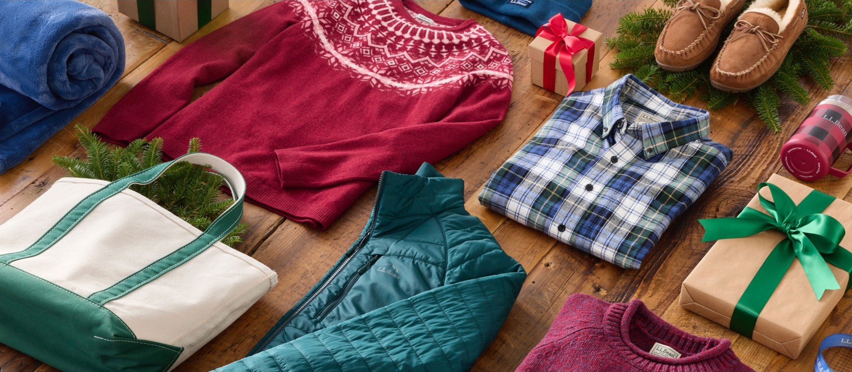 Winter clothes, wrapped gifts, a white tote bag, and pine branches arranged on a wooden floor in a festive holiday display.