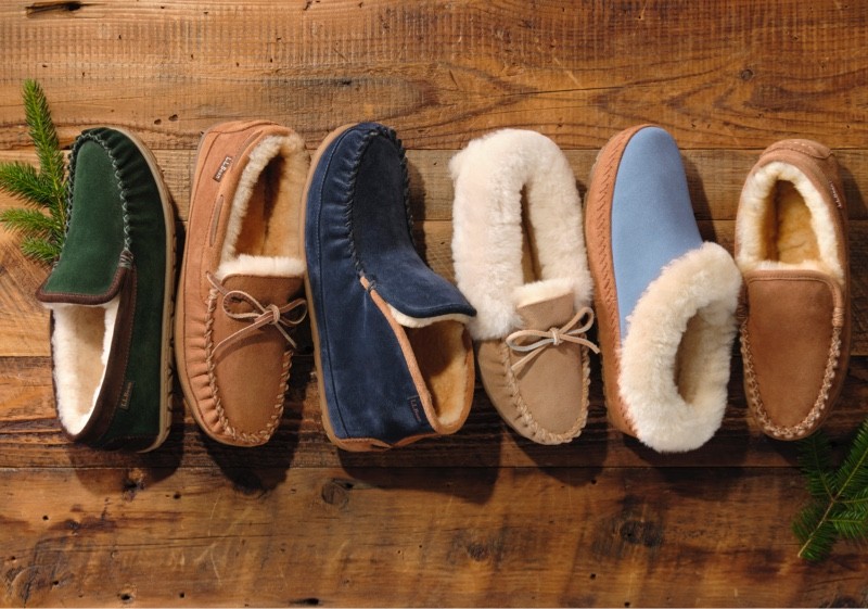 Five pairs of fur-lined moccasin slippers in assorted colors arranged on a wooden surface.
