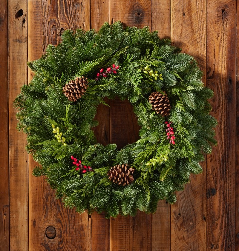 Green wreath with pinecones and red berries hanging on a wooden wall.