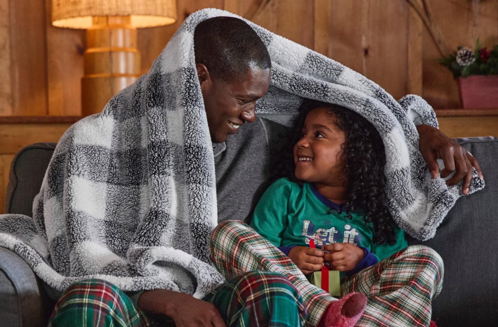 Two people in pajamas sit on a couch, sharing a checkered, cozy, plush fleece throw, with a lamp and wooden wall behind them.