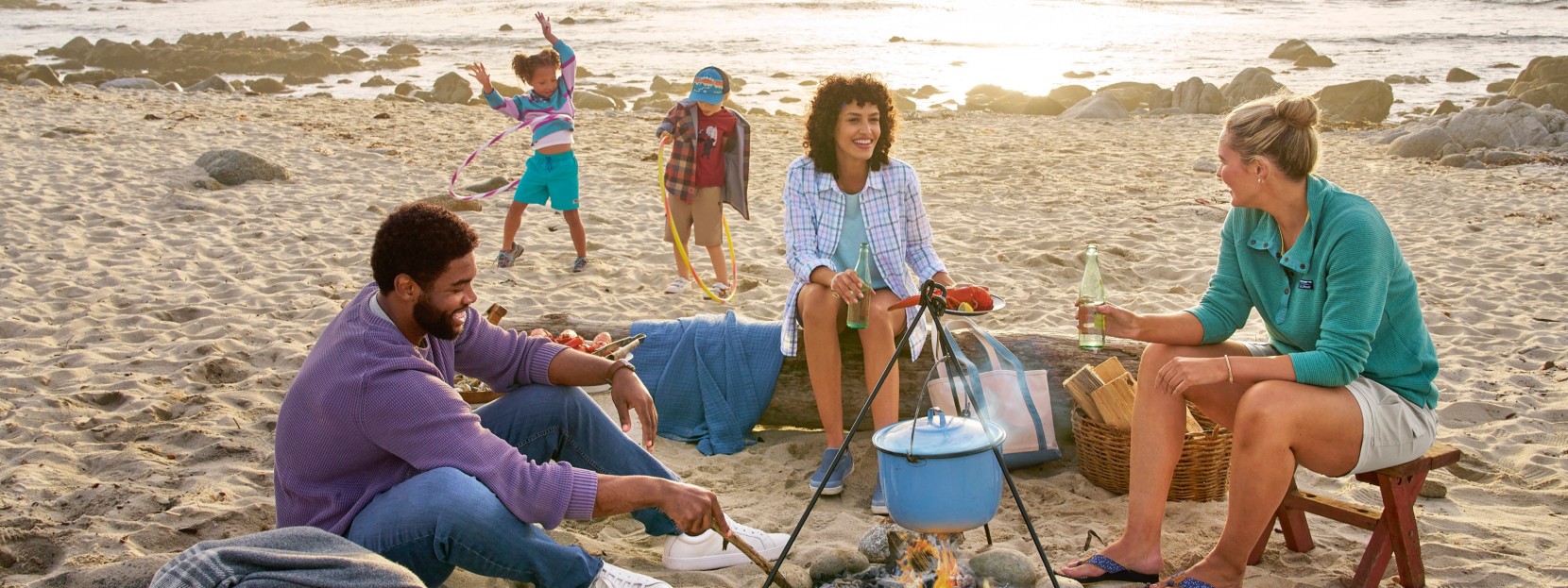 A happy group sits around a beach campfire with a hanging pot, while two kids play near the water.