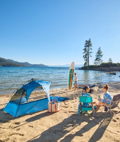 Two people relax in beach chairs by a blue sun shelter at a lake while a third person uses a stand up paddle board.