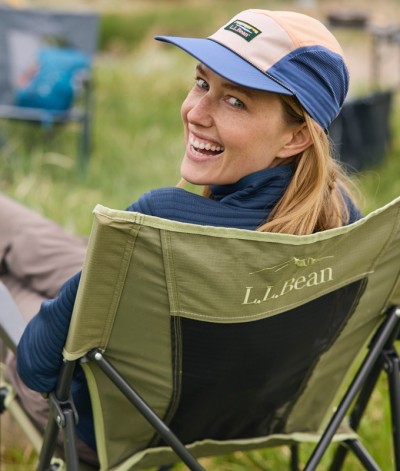Smiling woman sitting outdoors in a green L. L. Bean camping chair, wearing a blue and white cap.
