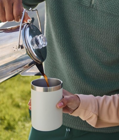 Coffee is poured from a stainless steel percolator into a white insulated camp mug held by another person.