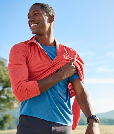 Smiling man in a blue shirt taking off a red jacket while standing outdoors in the sun.