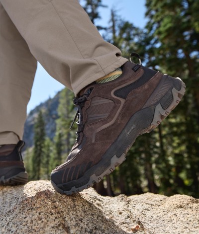 Hiking boot on a boulder with tan pants, set against a scenic forested mountain backdrop.