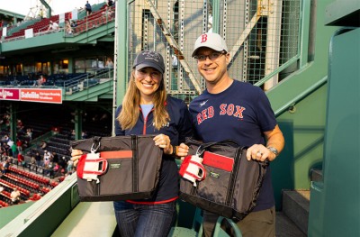 Two people in Red Sox gear smiling and holding black tote bags at Fenway stadium.