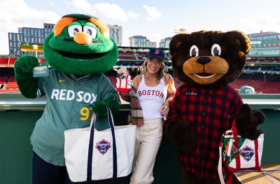 Person in Boston tank top with Red Sox cap stands between Wally and L.L. Bear mascots at Fenway Park, holding tote bags.