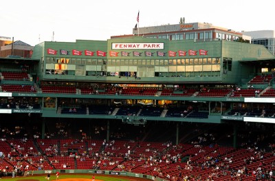 Fenway Park exterior with green facade, red banners, and “FENWAY PARK” sign above seating areas with scattered spectators.