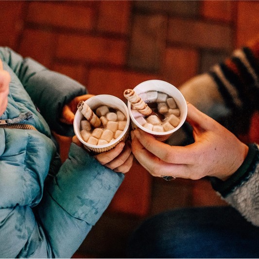Two people in winter clothes holding hot chocolate topped with marshmallows and wafer sticks.