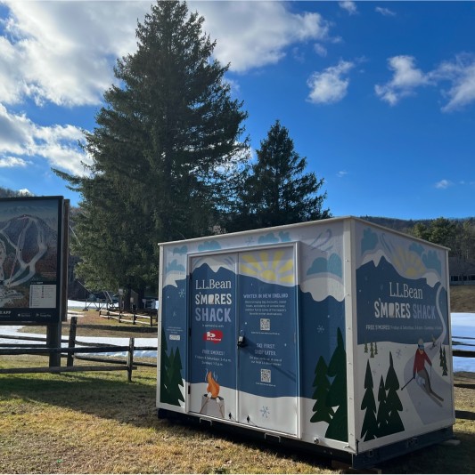 L. L. Bean S’mores Shack sits outdoors near tall pine trees under a bright blue sky with scattered clouds.