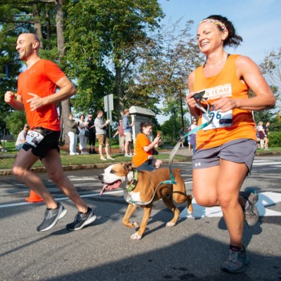 Two runners in orange shirts and a dog race past spectators during an outdoor event.