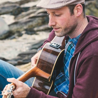 The performer Pete Kilpatrick poses for the camera, strumming a guitar on rocks.
