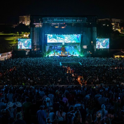  An outdoor concert at night from a distance with bright blue and green lights glowing from the stage.