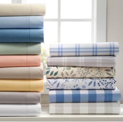 A stack of neatly folded, colorful bed sheets sit in front of a sunny window.