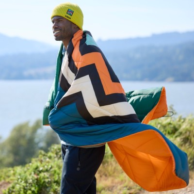 A man wrapped in a colorful, zigzag-patterned blanket stands outdoors near a body of water.