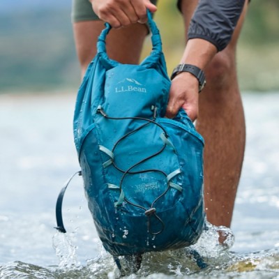 A person holding a blue L L Bean backpack in a stream, with water splashing around.