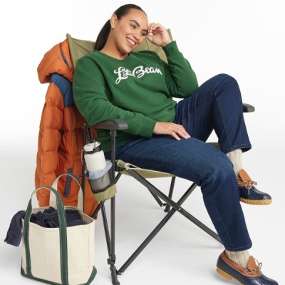 A woman sits in a foldable camping chair, wearing a green L L Bean sweatshirt, blue jeans, and casual shoes.