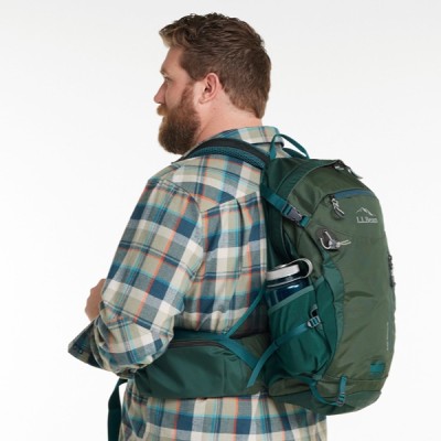 A man wearing a green backpack with a water bottle in the side pocket, dressed in a plaid shirt, facing away from the camera.