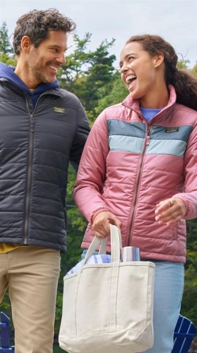 Two people outdoors wearing insulated jackets, carrying a tote bag with drink containers against a backdrop of green trees.
