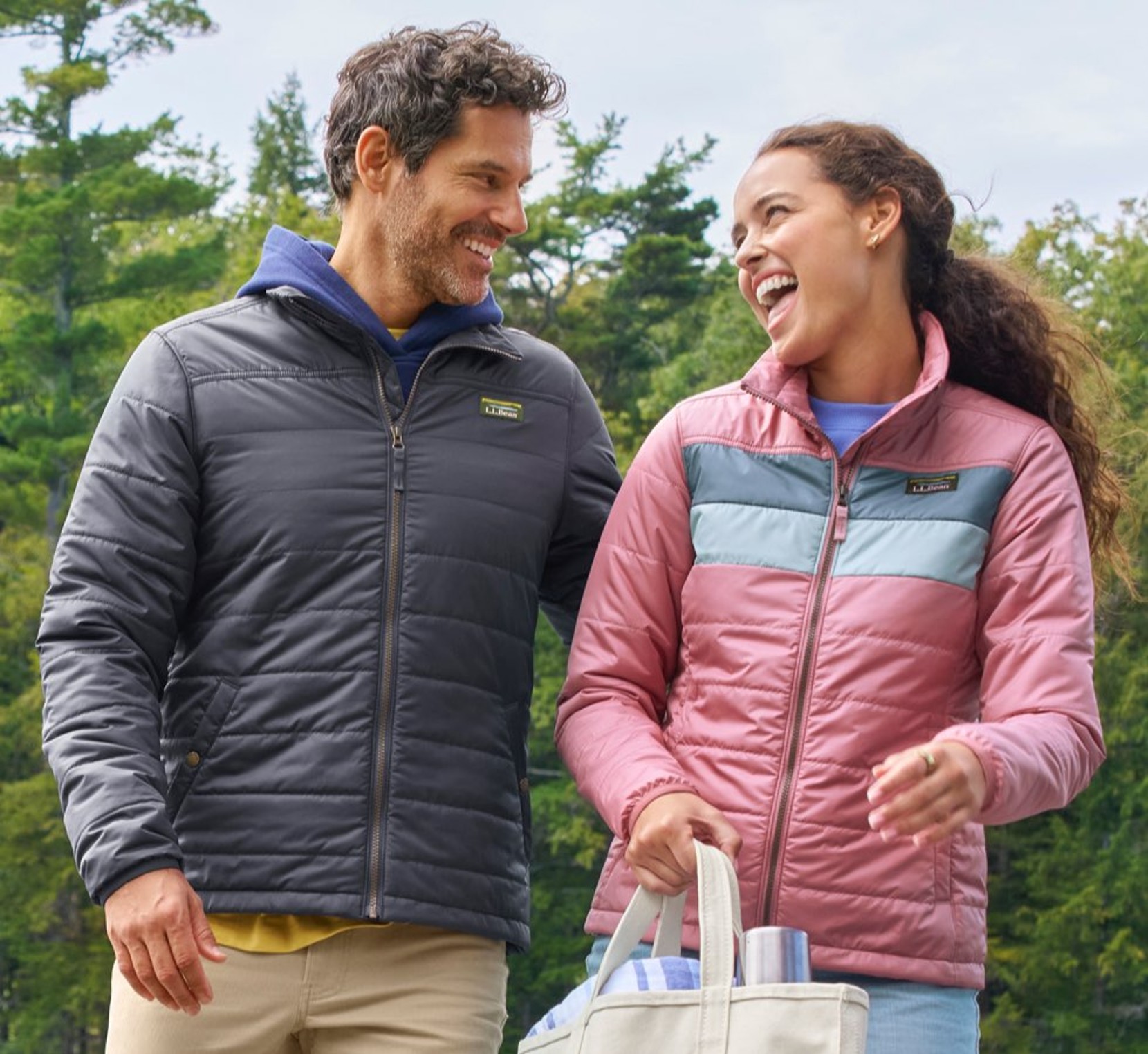 Two people outdoors wearing insulated jackets, carrying a tote bag with drink containers against a backdrop of green trees.