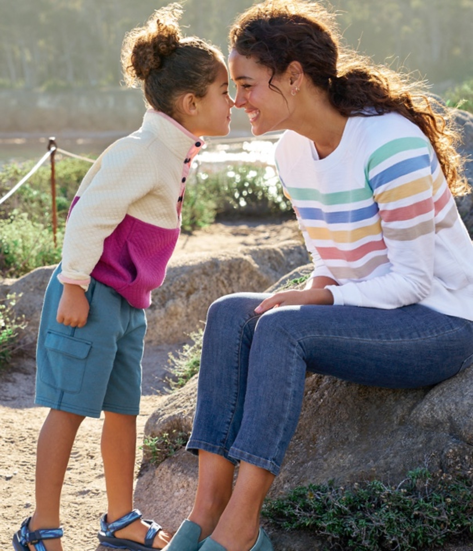 A child and an adult sit outdoors on a rock. The child leans in towards the adult, who is seated.