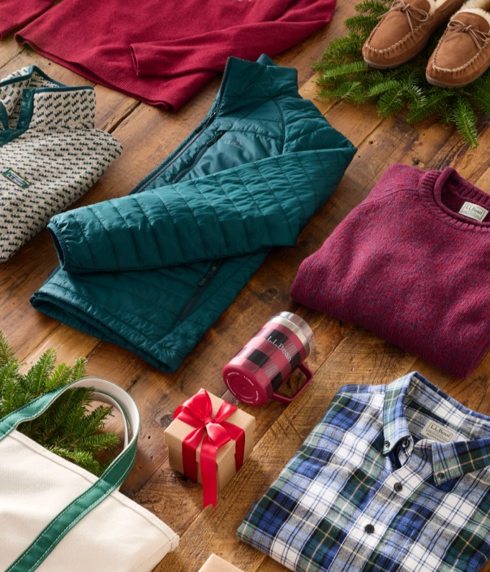 Winter clothes, wrapped gifts, a white tote bag, and pine branches arranged on a wooden floor in a festive holiday display.